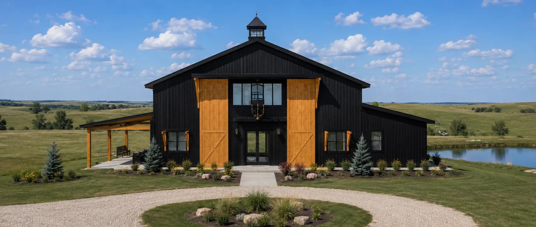 Architectural rendering of the front of Antelope Creek Ranch Venue near Wichita, Kansas: a black board-and-batten barn with two cedar sliding doors and a cupola, a covered porch on the left wing, and a gravel drive circling a planted island, with water and rolling prairie to the right.