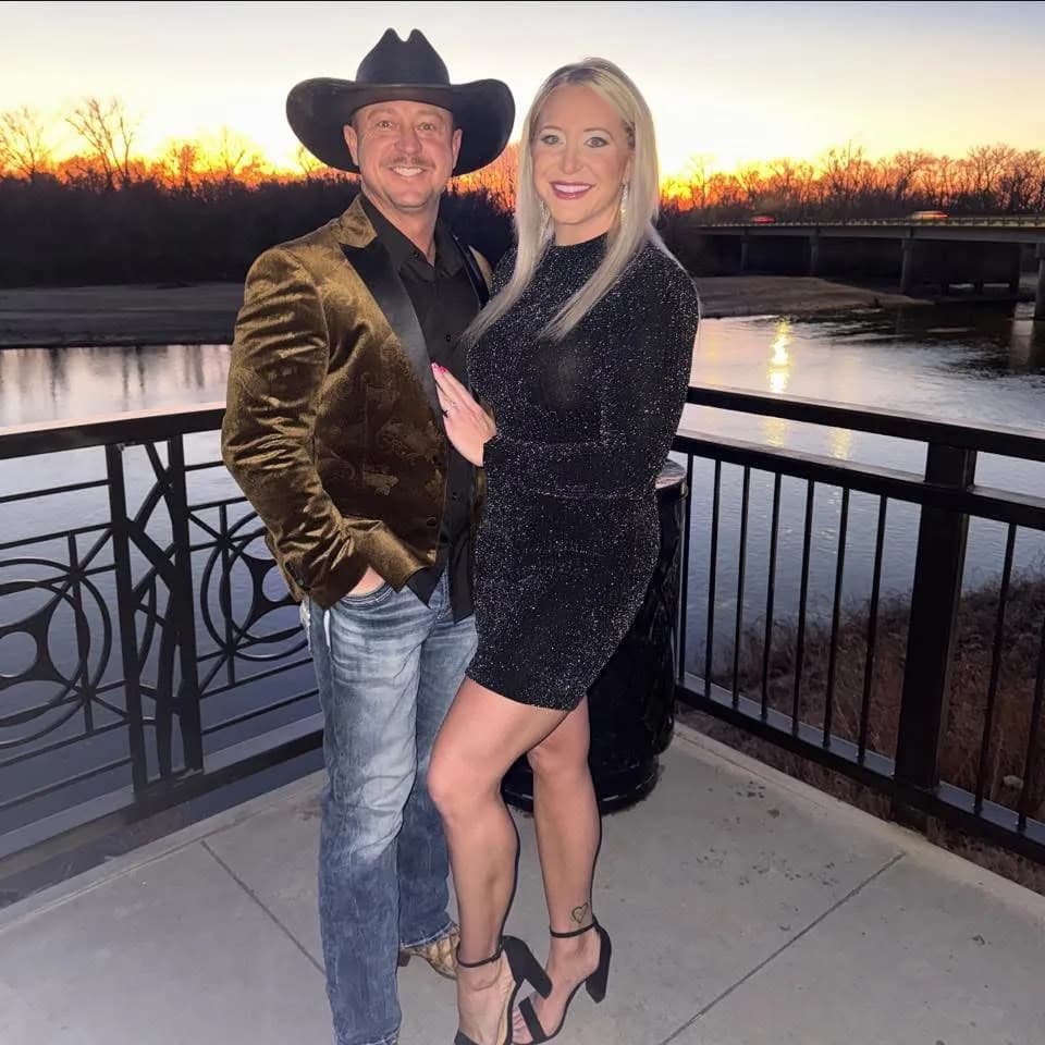 Debra and Michael Russell, the owners of Antelope Creek Ranch Venue near Wichita, Kansas, standing together at sunset on a railed walkway above the water, the treeline lit orange behind them.